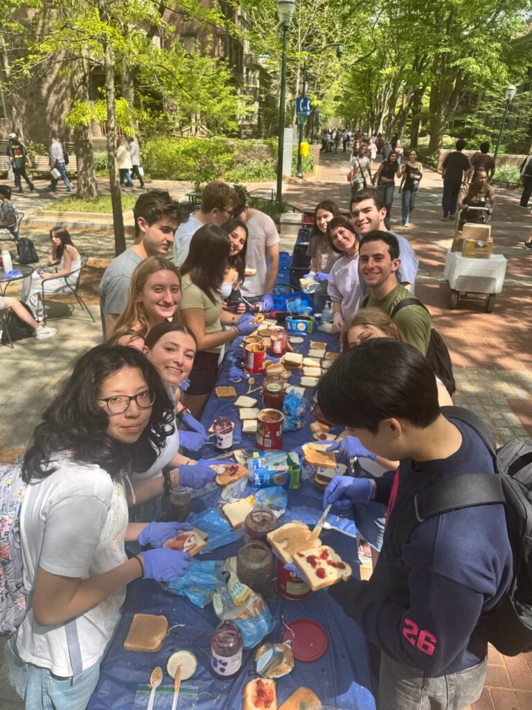 Penn students making peanut butter and jelly sandwiches at long tables outdoors on Locust Walk during the JHP PB&J-A-Thon